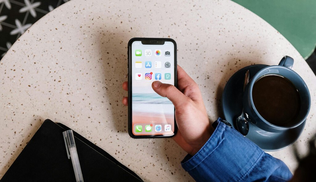 Hand holding iPhone with social media icons. Hand is over the top of a pink speckled table with deep blue coffee cup on.