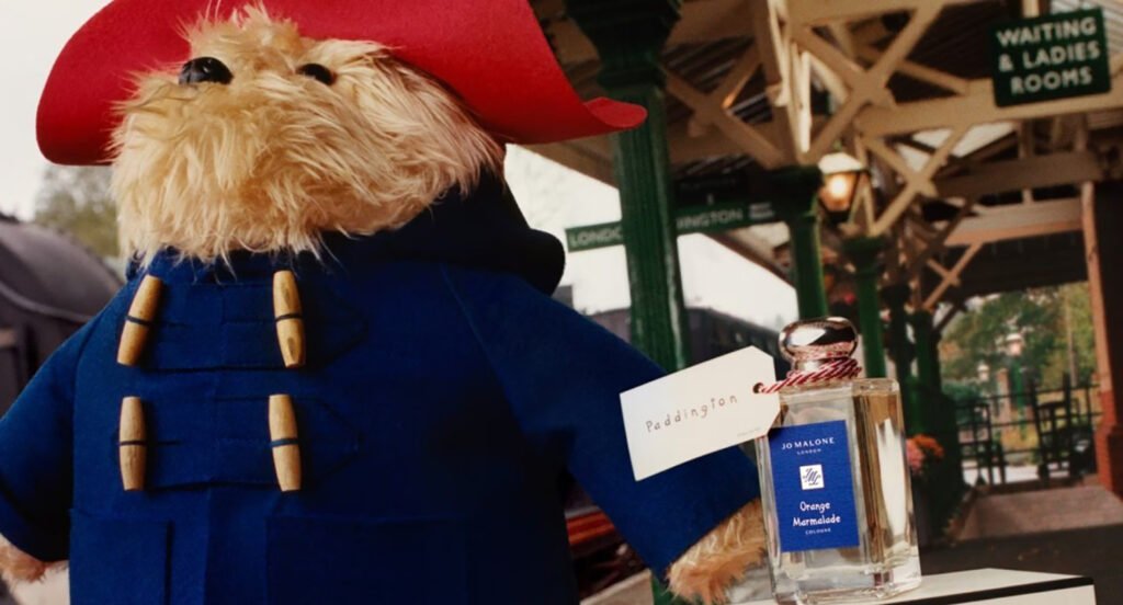 Paddington bear stuffed toy at a train station platform with a bottle of Jo Malone perfume next to him.