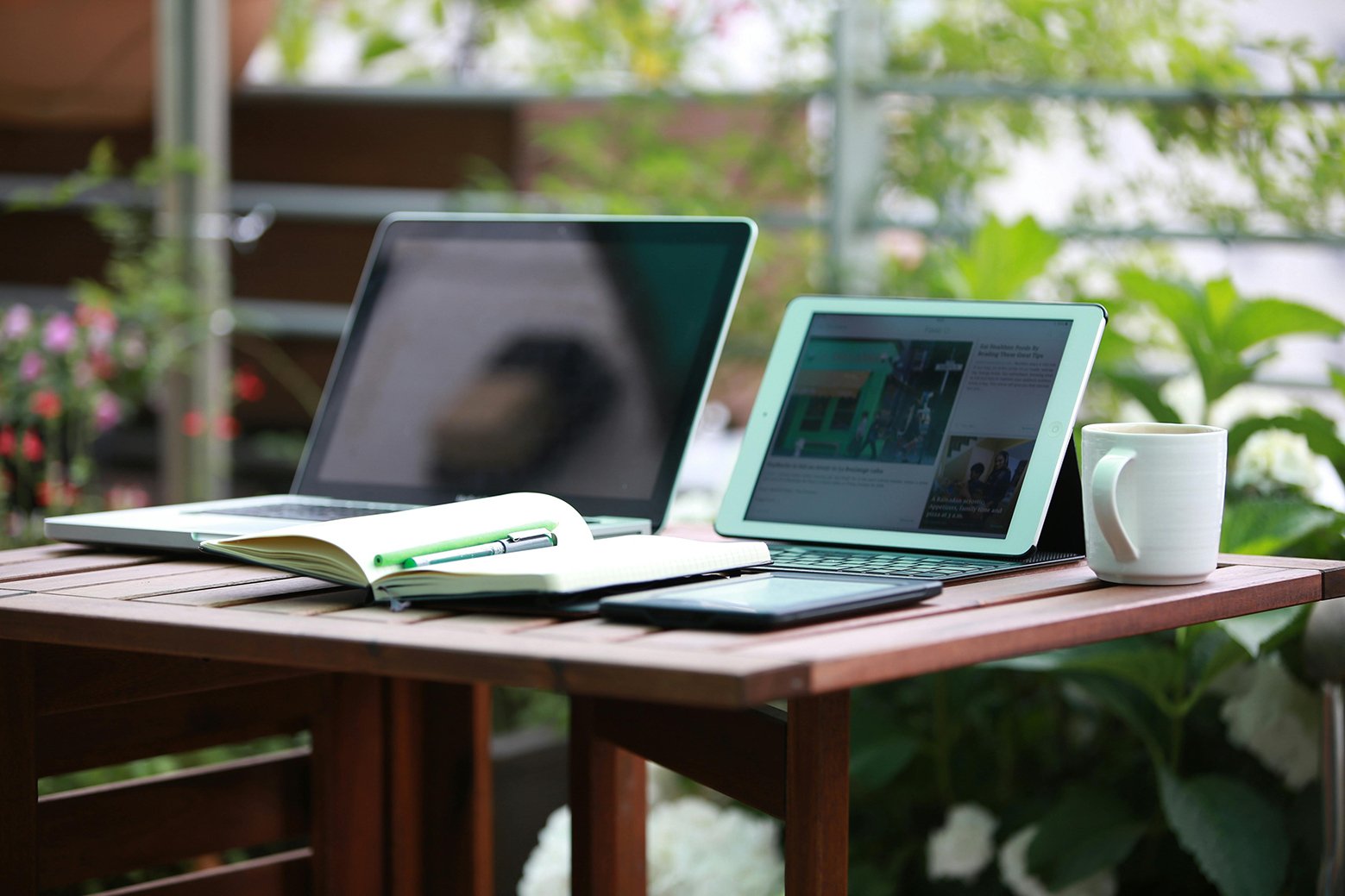 An open laptop and an iPad with a keyboard attachment sit on top of a wooden desk. A notepad is sprawled across the front of the desk. The picture shows that to know the most recent Instagram updates you should always be watching out for new news on different devices.