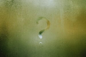 Question mark symbol drawn onto a foggy window with green poking through. Circular droplets of water are around the question mark.