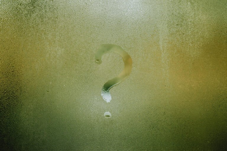 Question mark symbol drawn onto a foggy window with green poking through. Circular droplets of water are around the question mark.