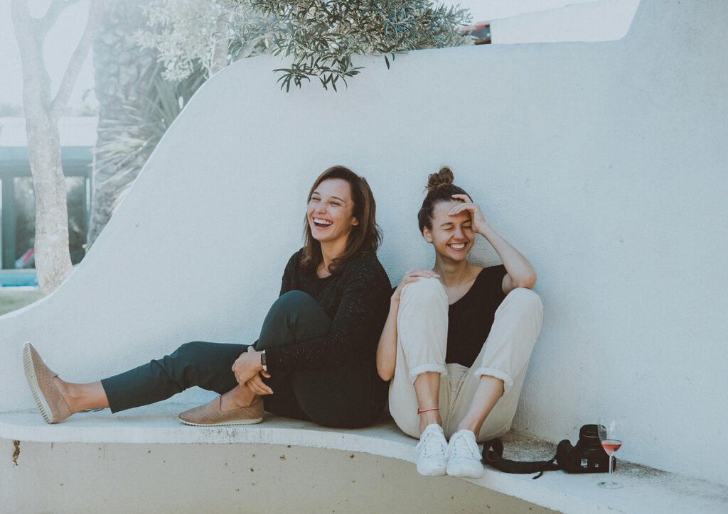 Two young women with brown hair, sat on a ledge laughing. There is a camera and a glass of wine next to them.