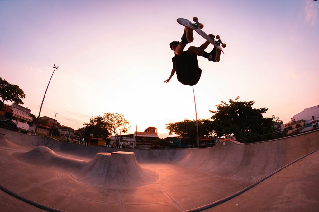 A skatepark at dusk with pink lighting. A skateboarder is doing a big trick and is high in the air above the skatepark.