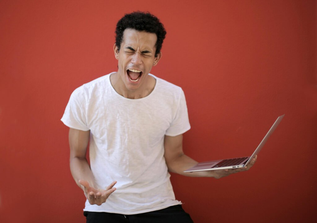 A man against a red background holding a laptop looking angry.