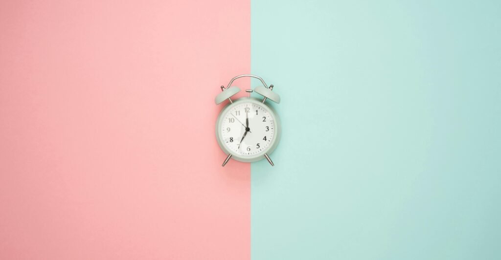 A duck egg blue alarm clock with silver hardware against half pastel pink and half pastel blue background. The clock shows the time as 7pm.