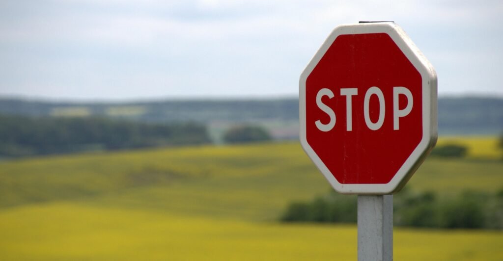 A red hexagonal stop sign on a grey steel pole. It is in focus against a fussy countryside background.