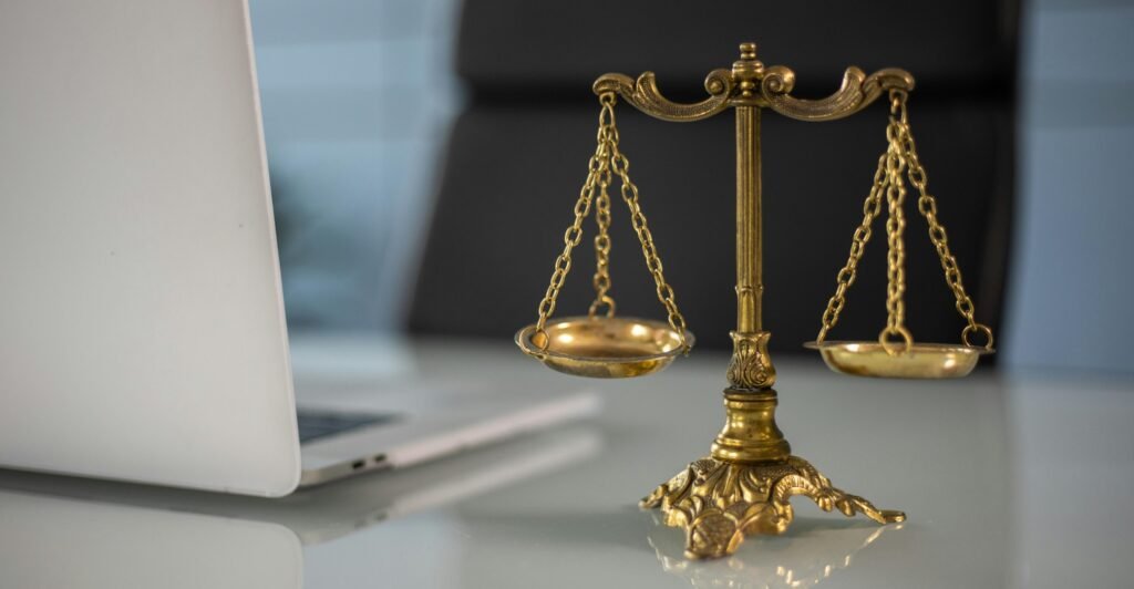 Gold scales of justice sit on top of a plain grey desk next to a sleek slate grey laptop. The scales are evenly weighed.