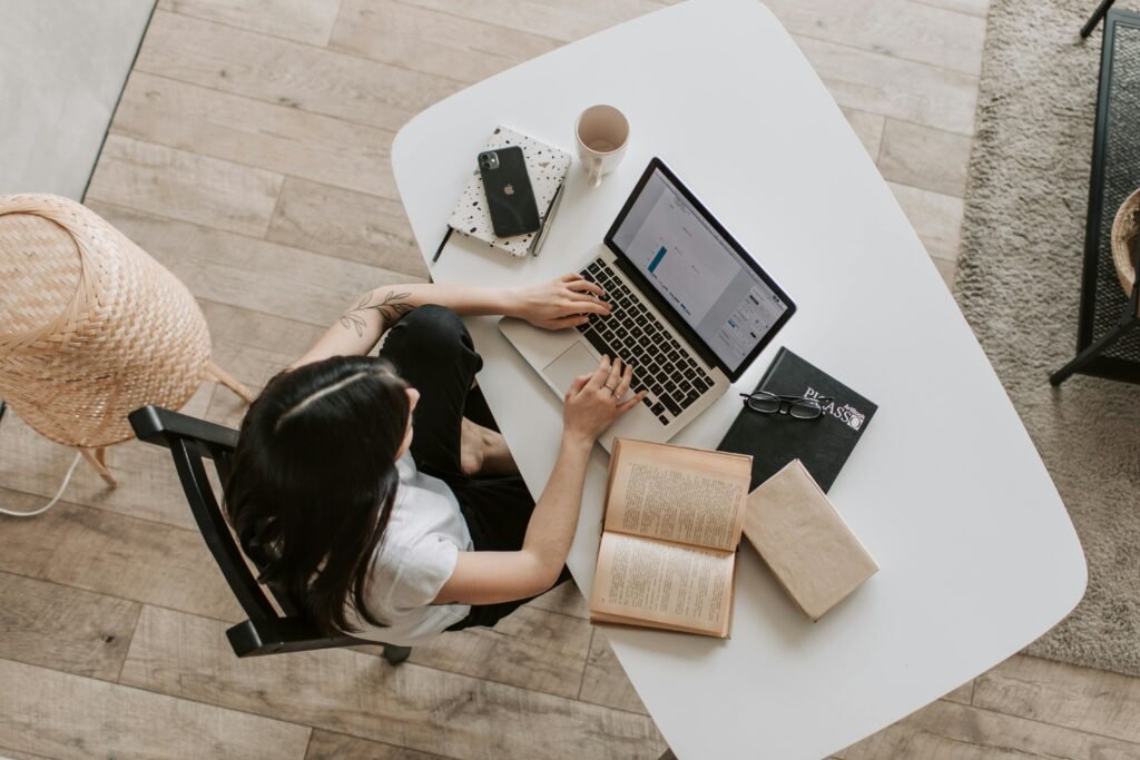 an image birds-eye view of a girl sat at a desk creating an email newsletter, also on the table are some notebooks, a book, an iPhone, an empty mug and a pair of glasses. The shot also includes a light wooden floor, and a freestanding wicker lamp. 