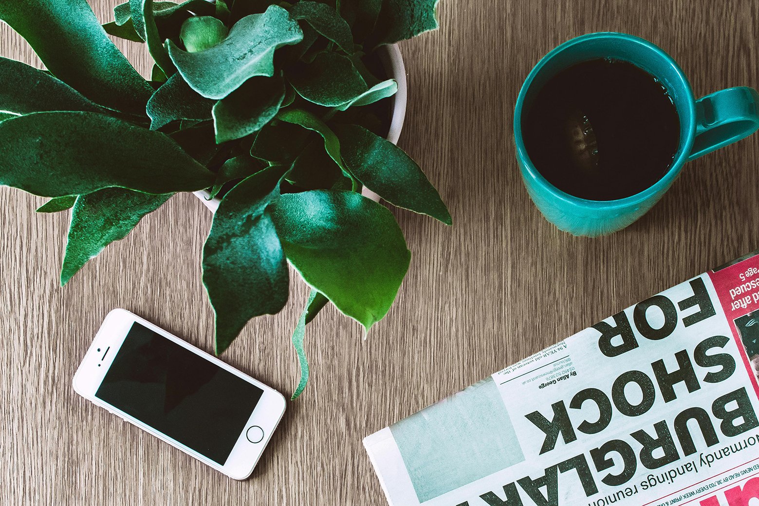 a birds-eye view of a wooden table with a plant, a mug of tea with the tea bag still in, an iPhone and a newspaper.