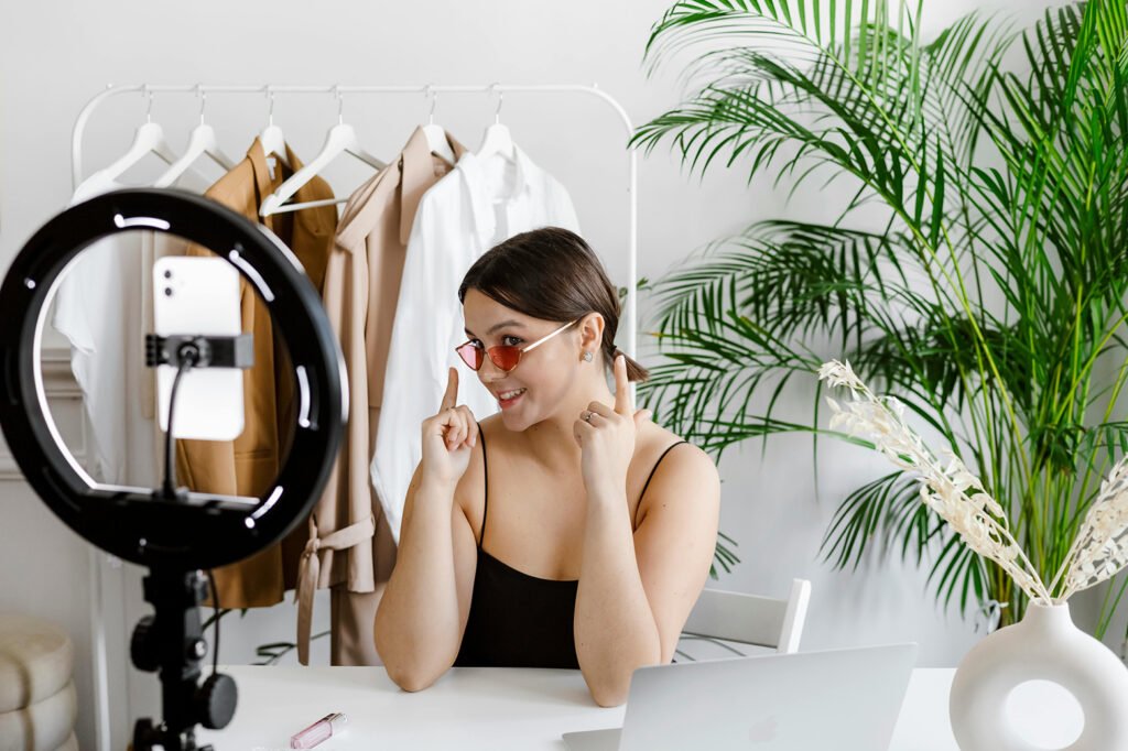 An image of a woman in front of a ring light with red glasses recording something on her phonw as she points to the glasses